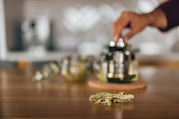 Focus on the dried herb on the table while the man uses a teapot in the background.