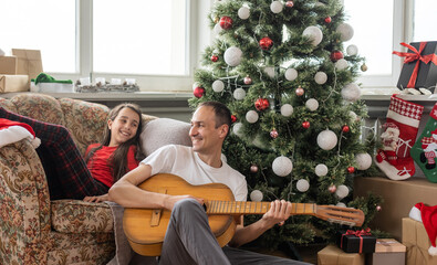 Father and little girl have fun together on Christmas holidays. Dad plays the guitar for his...