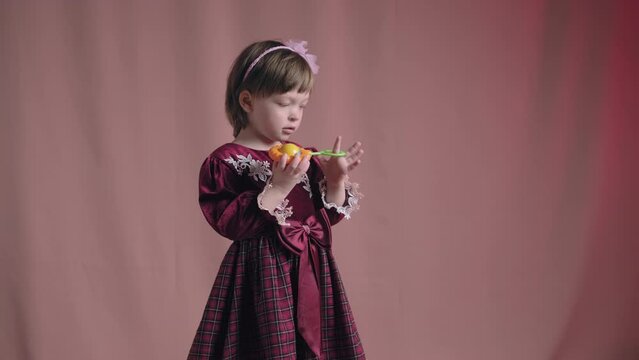 Cute little girl in an vintage burgundy dress is playing with a rattle toy. The concept of children theater productions and developing acting circles. Pink background
