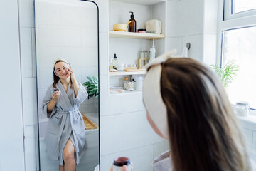 Young woman in bathrobe looking in the mirror and applying facial natural cosmetic clay mask on her...