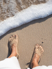 Man feet on sandy beach shore top view. A wave is coming to wet them.