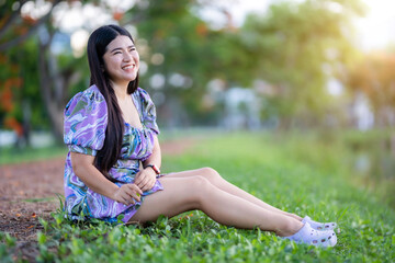 Happy Relaxing Portrait asian woman Wear purple dress wearing smartwatch while sitting on green grass lawn beside a reservoir at the city park outdoors.