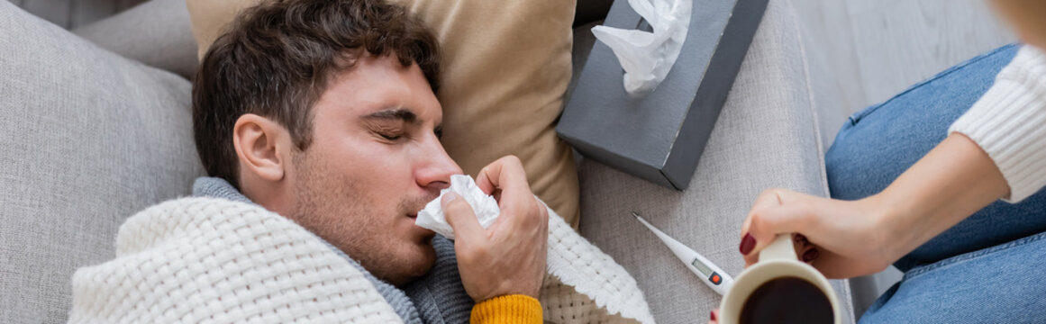 Top View Of Woman Holding Cup Of Tea Near Sick Boyfriend Lying Near Tissue Box, Banner.