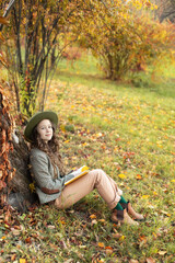 Young girl in hat and autumn clothes sit on lawn with  golden fall leaves. Happy girl in green hat and plaid jacket reading book on an autumn background. A smiling girl rest in fall park.