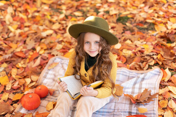 Young kid girl sit on plaid with pumpkins and reading book. Happy little girl in hat and autumn clothes on an autumn background. A smiling child is playing in fall park. 