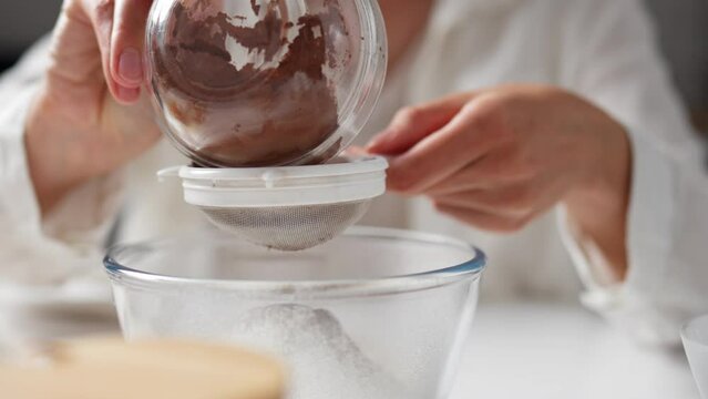 Woman in kitchen is sifting cocoa powder into  flour. Woman's hand holding sieve pours, sifts cocoa into bowl. Prepare dough for delicious homemade pie. Preparing ingredients for pie
