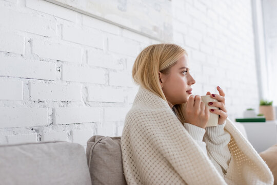 young woman in sweater sitting covered in blanket with cup of tea in living room.