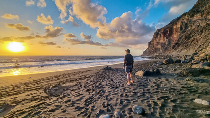 Man with hat walking barefoot on volcanic sand beach Playa del Ingles during sunset in Valle Gran...