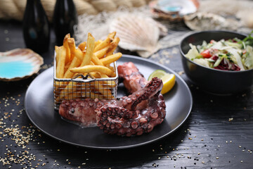 Grilled octopus tentacles served with potato fries in metal serving basket, on a black plate. Lunch set in a restaurant.