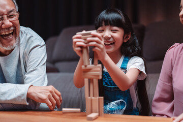 Asian happy family of little daughter play toy wooden block in home. Grandparents and young woman loving couple parent having fun play stack tower wood block game at night in living room.