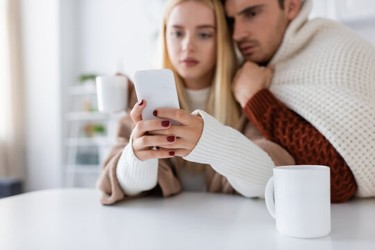 blurred woman using smartphone near boyfriend and focused cup of tea on table.