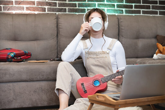 Cozy young hipster woman in casual wear drinks hot coffee from white cup, takes a break between learning to play ukulele guitar, sitting on floor in living room at home, leisure concept
