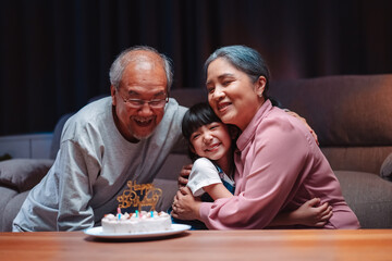 Asian happy family of little girl hugging lover with grandparents after blowing out candles on cake. Celebrate birthday anniversary party on table at night in living room, having happiness lifestyle.