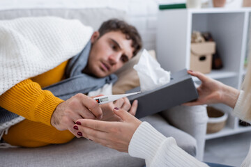 caring girlfriend holding tissue box and digital thermometer near sick boyfriend lying on sofa...