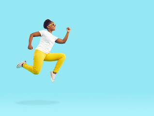 Side view happy energetic young African American woman in white T shirt, yellow trousers and sunglasses running, flying or jumping high in air isolated on bright blue blank empty copy space background