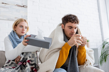 blonde woman in scarf holding tissue box near sick boyfriend with cup of tea.