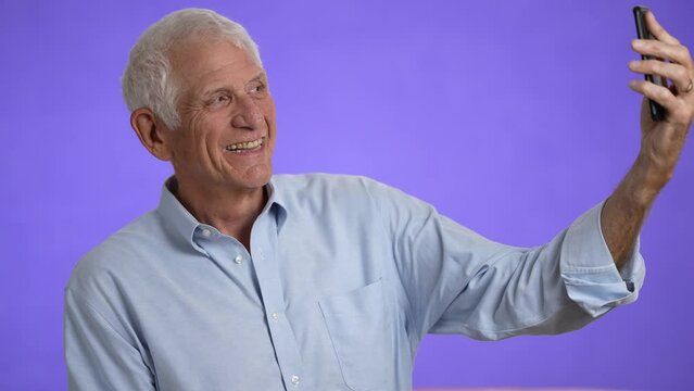 Happy elderly gray-haired man 70s in blue shirt doing selfie shot mobile phone post photo on social network isolated on plain light purple background studio portrait