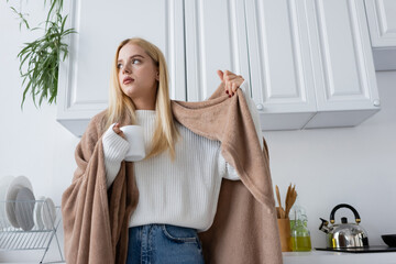 low angle view of young blonde woman in white sweater and blanket holding cup of tea.