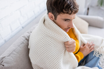 high angle view of young man covered in blanket sitting on sofa and getting warm at home.