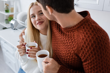 cheerful young couple in knitted sweaters hugging and holding cups with tea while warming up in winter.