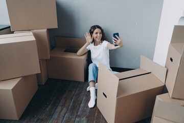 Happy girl tenant homeowner chatting via video call sitting on floor near carton boxes on moving day