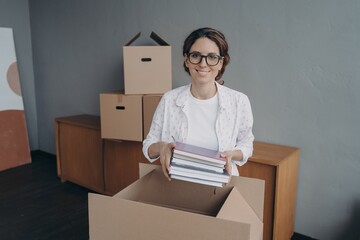 Smiling hispanic female office worker in glasses packs things in carton box preparing for relocation