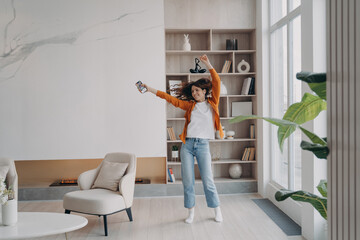 Energetic woman dancing in living room by music from smartphone in modern apartment home interior - obrazy, fototapety, plakaty