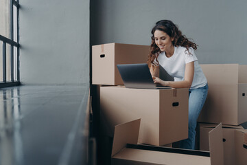 Female with boxes works at laptop, orders moving company, shopping online. Ecommerce, relocation day
