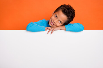 Photo portrait of adorable small pupil boy hide behind blank ad print space dressed stylish blue look isolated on orange color background
