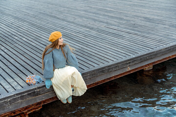 Outdoors fashion portrait of a beautiful middle aged woman walking on the beach. Marine background. Dressed in a stylish warm blue sweater, yellow skirt and beret.