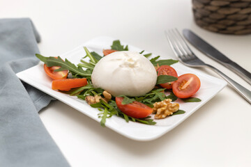 Fresh burrata cheese with arugula leaves, cherry tomatoes and walnuts on a white serving plate on a white table. Traditional Italian cheese