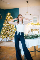 Attractive young girl in white blouse and trendy trousers  with hairstyle and make-up in room decorated for Christmas by the holiday table. Festive mood. Christmas