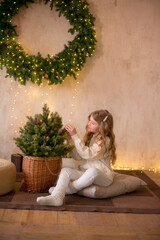 Obraz premium Little cute girl with blond long hair in a warm knitted sweater by a Christmas tree and a big Christmas wreath and burning garlands. Christmas mood 