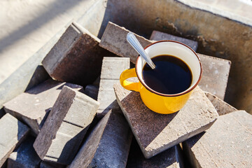 Fototapeta na wymiar A cup of coffee at a construction site. A drink in a yellow mug is located on a pile of paving stones. Rest during the working day. Close-up