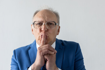 Portrait of serious mature businessman with finger on lips. Close-up of senior manager wearing formalwear and eyeglasses looking at camera and making silence gesture. Secret and silence concept