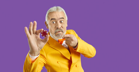 Portrait of crazy goofy bearded adult man in funky yellow suit on purple studio background pointing at poker chip and looking at it with funny angry face expression. Casino, gambling addiction concept - obrazy, fototapety, plakaty
