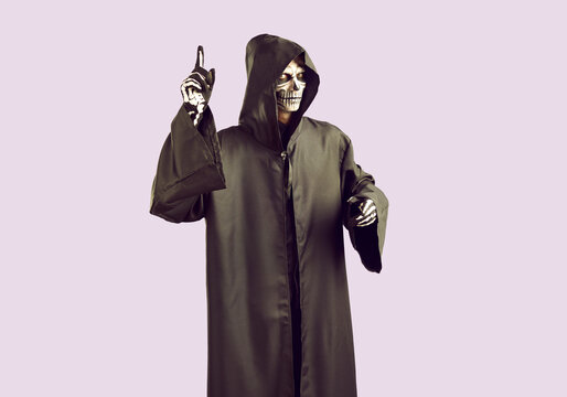 Man in image of angel of death shows finger up with idea for Halloween party celebration. Young man with creepy skeleton makeup and in long black cloak with hood poses against light lilac background.