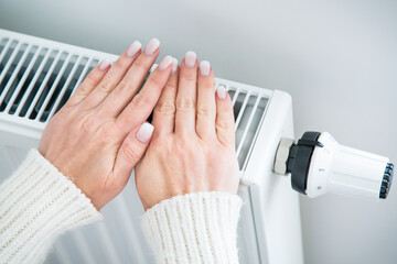 Woman heating her hands near the radiator, cold in the house. Saving heating. Expensive utility bills.