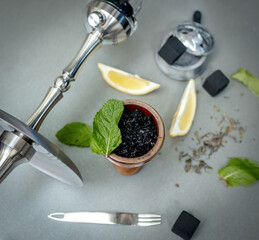 Hookah bowl with aromatic tobacco with lemon and mint on gray table with smoking accessories. Hookah smoking in a hookah lounge. Steel hookah, tongs, tea and coconut charcoal