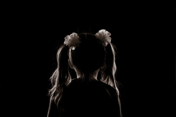 silhouette of a girl with two ponytails sitting with her back in the dark. anonymity and child...