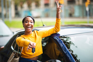 Happy black woman leaning on her new car and showing the keys with peace hand sign