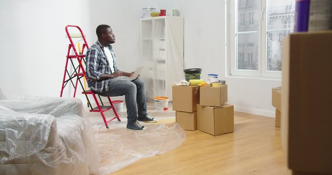 Serious African American young male interior designer sitting on red ladder in empty apartment with boxes typing on laptop searching internet choosing new decor for room, renovation, repair concept
