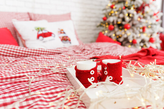 red knitted covers stand on a tray on a bed with a checkered bed against the background of white pillows with a New Year's pattern of a bright Christmas tree with flickering lights