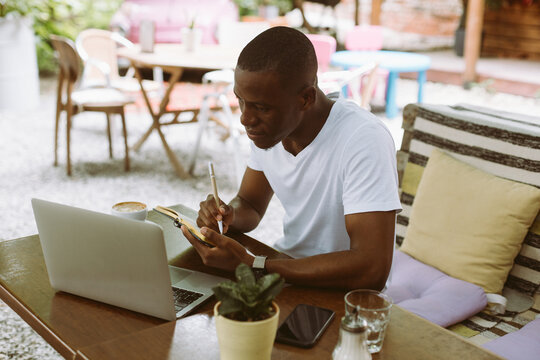  concentrated modern dark skin man in casual clothes writing business plan in textbook during webinar on laptop in outdoor coffee shop. Businessman sitting with smartphone and PC