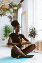Vertical calm and spiritual young dark skin woman with close eyes, join hands in namaste Asana pose, sitting in lotus position. Yoga practicing at home. Healthy lifestyle. Relaxing and rest on weekend
