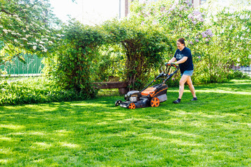 Brown-haired female gardener in casual clothes using gasoline lawn mower in the yard, grounds....
