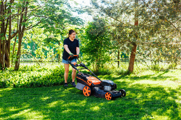 Tired brown-haired woman in casual clothes using gasoline lawn mower or grass cutter, caring her lawn with organic living plants in countryside. Cutting fresh lush green grass in sunny day in backyard