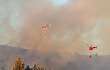 Two fire-fighting helicopters carry water to extinguish a fire on Mount San Cristóbal in Pamplona. Intense dark smoke covers the sky and helicopters.