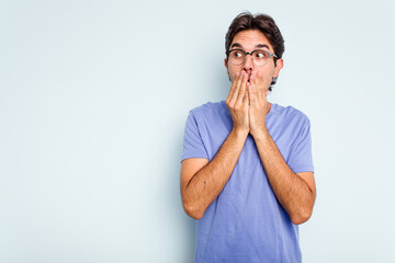Young hispanic man isolated on blue background thoughtful looking to a copy space covering mouth with hand.