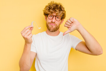 Young caucasian man wearing hearing aid isolated on yellow background showing a dislike gesture,...